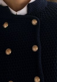 Granatowy, strukturalny płaszcz z złotymi guzikami, podwójnym zapięciem i wysokim kołnierzem. Tkanina ma wzór tkany.