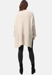 Beige Strickjacke mit lockerer Passform, langen Ärmeln und asymmetrischem Saum. Sie hat eine strukturierte Maschenoptik und seitliche Schlitze für Bewegungsfreiheit.