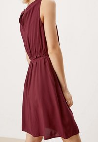 Vestido sin mangas color marrón con una cintura ajustada, tejido suave y falda suelta. Presenta un detalle de lazo lateral y un respaldo fruncido.