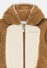 Veste en polaire marron avec un devant crème, dotée d'une fermeture éclair et d'une capuche duveteuse. Texture douce, design léger et couleurs contrastantes.
