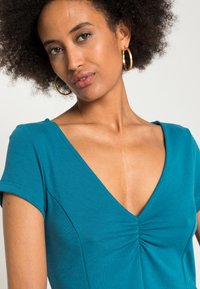 Top em verde-água com decote em V e mangas curtas, apresentando um detalhe franzido no centro. Textura de tecido suave, projetado para ajustar-se de perto.