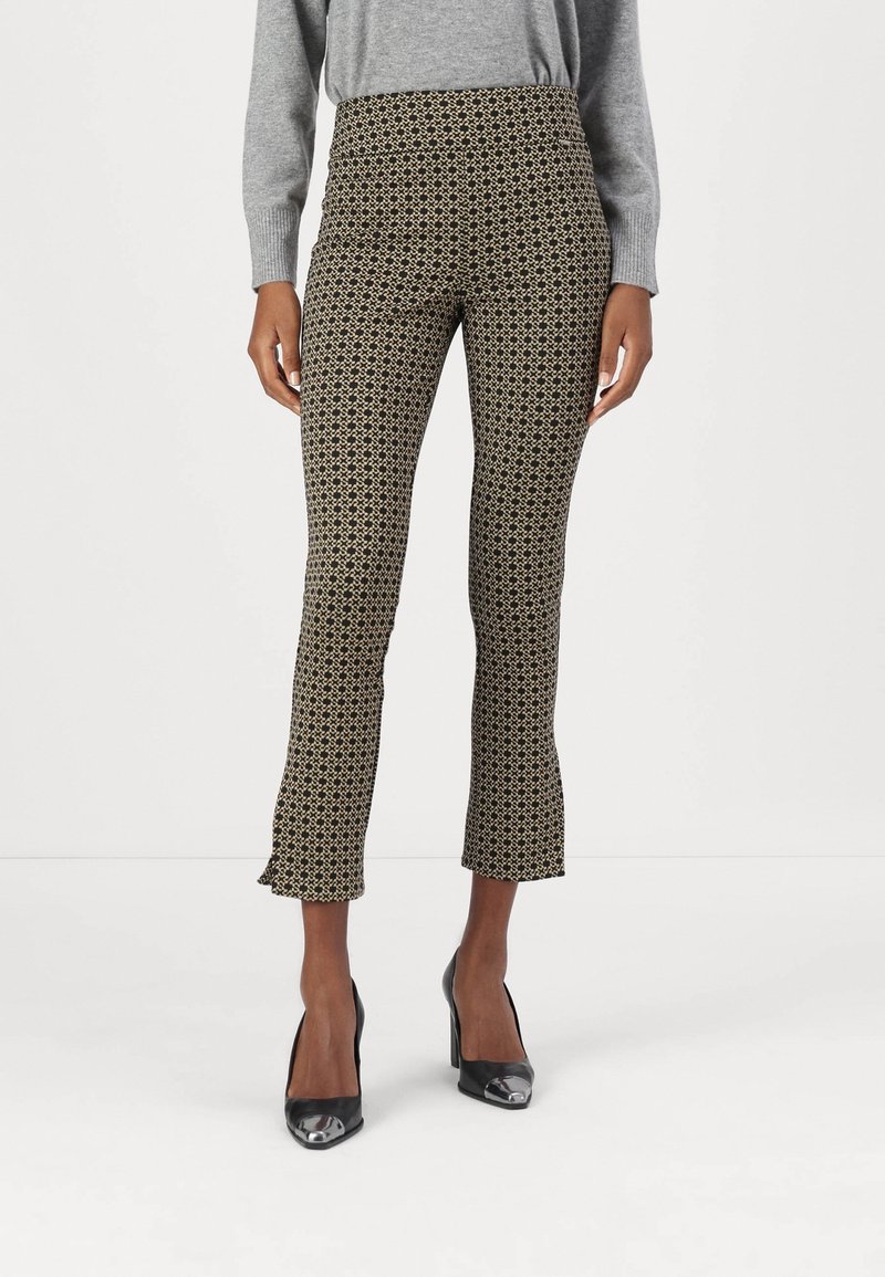 Femme portant un pantalon court taille haute avec un motif géométrique noir et beige, un pull gris et des chaussures à talons noirs.