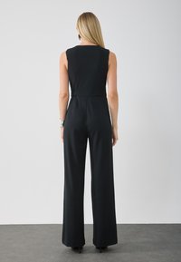 Musta ilmanhiha jumpsuit, jossa on muotoiltu yläosa ja leveät lahkeet. Valmistettu sileästä kankaasta, jossa on korkea kaulus ja sivutaskut.