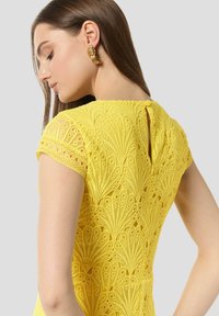 Gelbes Spitzenkleid mit Blumendesign, kurzen Ärmeln, einem Schlüssellochverschluss auf der Rückseite und strukturiertem Stoff. Goldene Creolen betonen den Look.