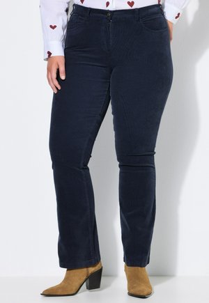 Persona que lleva pantalones ajustados azul oscuro, una camisa blanca con patrones de corazones y botas de ante marrón claro con tacón, de pie contra una pared clara.