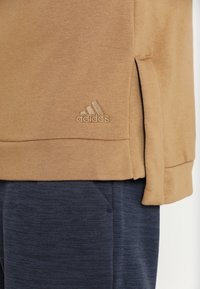 Sudadera marrón con slit laterales, hecha de un suave material de algodón. Presenta un logotipo bordado de Adidas en un tono a juego.