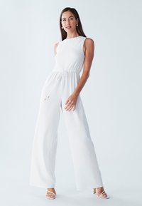Witte mouwloze jumpsuit met een gestructureerde corsage, brede pijpen, verstelbare strik taille en subtiele gouden hardware accenten.