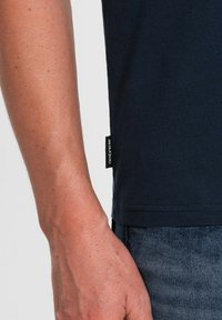 Námořnické bavlněné tričko zobrazené s detailním záběrem na rukáv a zakončení, s malou tkanou nášivkou s nápisem "ombre".