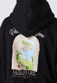 Sweat à capuche noir avec une illustration d'une arche représentant une ville côtière, des bateaux, de la verdure et le texte "Luxury Life Dreaming of Monaco."