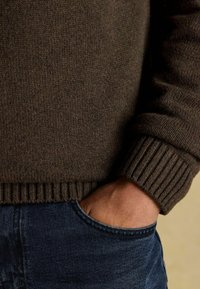 Brauner gestrickter Pullover mit gerippten Bündchen und Saum, kombiniert mit dunkelblauen Jeans. Nahaufnahme, die Textur und Farbdetails zeigt.