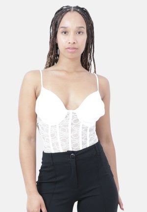 Femme aux longues tresses portant un corset blanc en dentelle à fines bretelles et un pantalon noir taille haute, debout devant un fond clair et uni.
