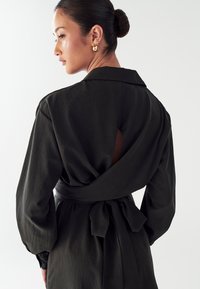 Must mustar, pikad varrukad ja krae, selja lõhiku ja seotava vööga. Tekstuuriga kangas omab peent läiget, mis lisab disainile sügavust.