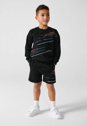 Junger Junge steht mit den Händen in den Taschen, trägt einen schwarzen Pullover und kurze Hosen mit mehrfarbigen Streifenmustern sowie weiße Turnschuhe.