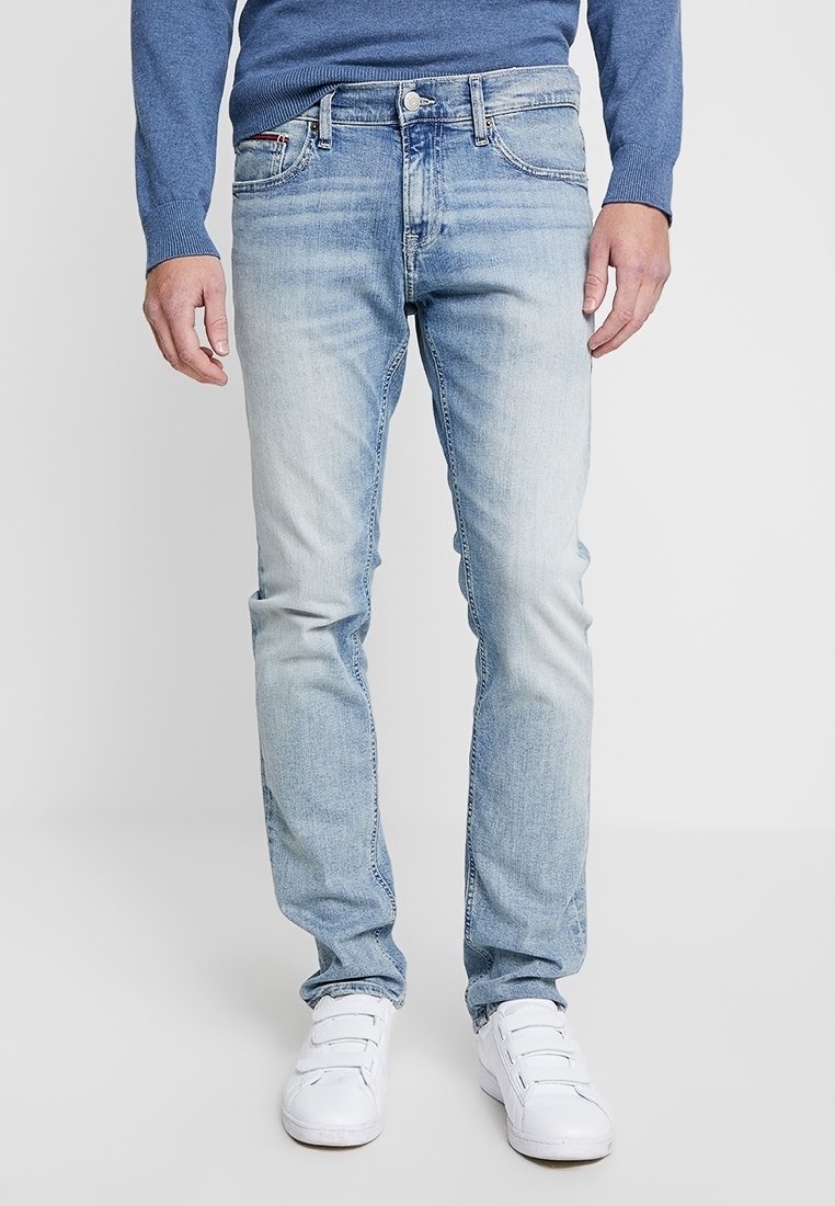 Man som bär ljusblå slim-fit jeans, blå tröja och vita velcro-sneakers, stående mot en enfärgad ljusgrå bakgrund.