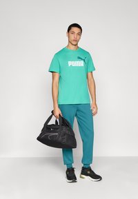 Τιρκουάζ μπλουζάκι με κοντά μανίκια και λευκό λογότυπο "PUMA", συνδυασμένο με βεραμάν παντελόνι φόρμας και μαύρα αθλητικά παπούτσια. Κρατάει μια μαύρη τσάντα duffel.