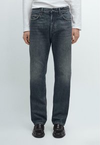 Jeans blu scuro con taglio dritto, leggermente sbiaditi, cinque tasche e dettagli in metallo; abbinati a scarpe nere.