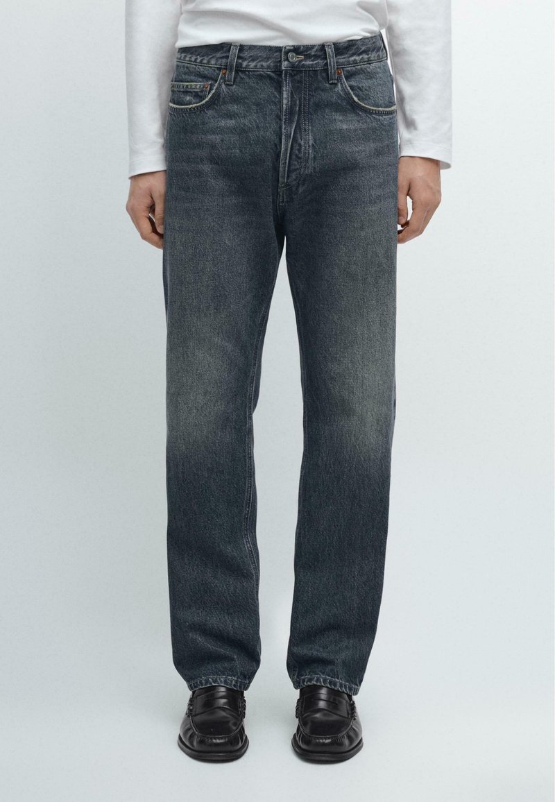 Jeans blu scuro con taglio dritto, leggermente sbiaditi, cinque tasche e dettagli in metallo; abbinati a scarpe nere.