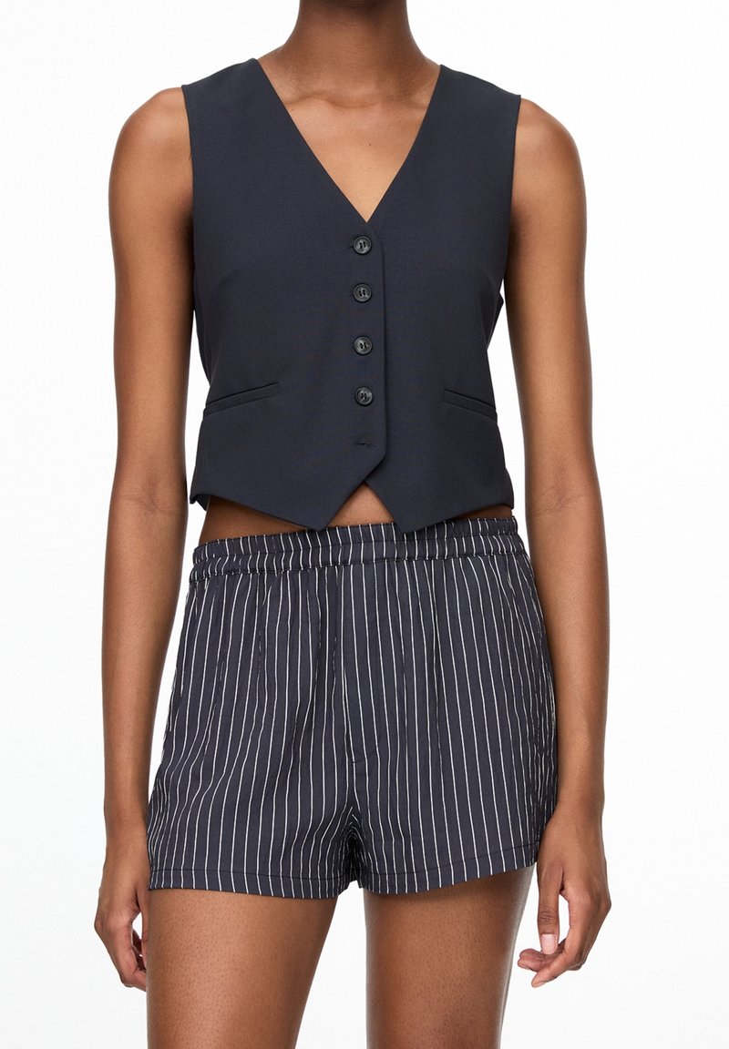 Femme portant un gilet sans manches noir boutonné et un short noir à fines rayures blanches verticales, debout les bras détendus.