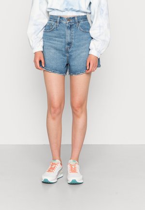 Persoon die hooggetailleerde blauwe denimshorts draagt, een lichtgekleurde top met lange mouwen en witte sneakers met perzikkleurige veters, staand tegen een effen achtergrond.