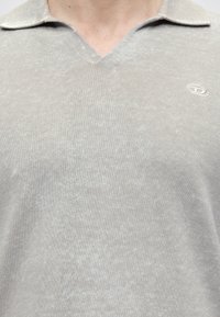 Gros plan d'une personne portant un polo en maille gris clair avec un petit logo ovale blanc sur le côté gauche de la poitrine.