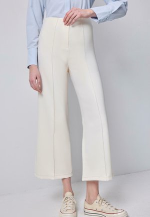 Femme portant un pantalon large taille haute couleur crème, une chemise à manches longues bleu clair, et des baskets crème à lacets, debout devant un fond uni.