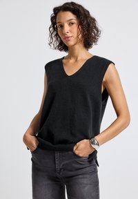 Kvinna med kort lockigt hår, iklädd ärmlös svart stickad topp och mörka jeans, med händerna i fickorna, stående mot en enfärgad ljus bakgrund.