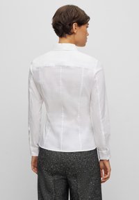 Camicia bianca a maniche lunghe con un design aderente, texture liscia e dettagli di cucitura sottili. Presenta un colletto classico e polsini con bottoni.