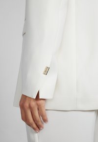 Valkoinen bleiserin hiha, jossa on istuva leikkaus, kultaiset metalliyksityiskohdat hihassa ja sileä kangas.