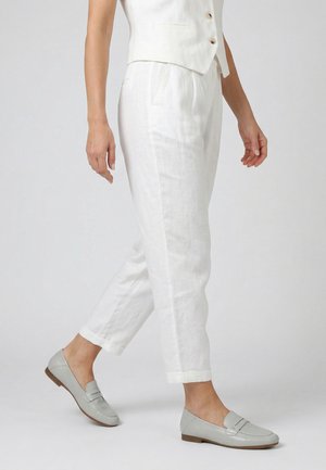 Kvinde gående iført hvide korte bukser, en beige vest og lysegrå flade loafers på en ensartet hvid baggrund.