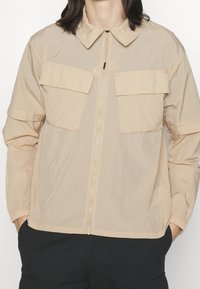 Giacca leggera beige con zip frontale e due tasche sul petto. Presenta un colletto classico e polsini elasticizzati, realizzata in un tessuto morbido.