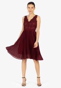 Robe sans manches bordeaux avec un corsage en dentelle, un décolleté en V, une jupe plissée et un tissu doux, se terminant au niveau du genou. Sandales à brides à la cheville noires.