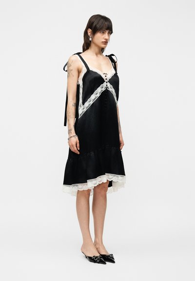 Γυναίκα με τατουάζ, που φορεί μαύρο σατέν φόρεμα μέχρι το γόνατο με λευκή δαντέλα στο τελείωμα, φερμουάρ δεμένο στους ώμους και μαύρες μυτερές γόβες.