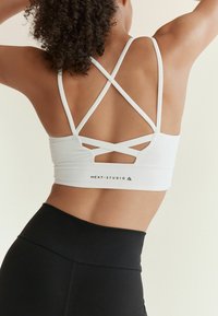 Witte sportbeha met kruisbanden op de rug en logo-detail aan de onderkant, gecombineerd met hoge zwarte leggings. Glad textuur.