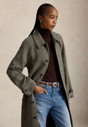 Femme aux cheveux tressés porte un long manteau à chevrons sur un col roulé sombre et un jean bleu avec une ceinture marron, debout avec une main dans la poche.