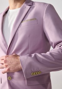 Lys lilla blazer laget av glatt stoff, med en lomme med beige detalj, og fire runde gule knapper på ermene.