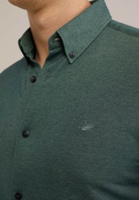 Camisa verde de botones, hecha de tela texturizada, con cuello puntiagudo, botones negros y un sutil logo bordado en el pecho.