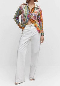 Chemise à manches longues colorée et à motifs avec un devant noué, assortie à un pantalon taille haute blanche à jambes larges et des talons ouverts.