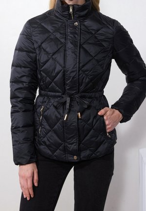 Kobieta w czarnej pikowanej kurtce z przednim zamkiem błyskawicznym, zatrzaskami, sznurkiem w talii, kieszeniami na zamek i wysokim kołnierzem, stojąca z jedną ręką w kieszeni.