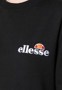 Sudadera negra con cuello acanalado, que presenta el logotipo bordado de "ellesse" en blanco y un acento en forma de medio círculo en rojo/naranja encima.