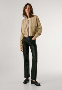 Giacca bomber cropped beige con due tasche frontali, abbinata a pantaloni in ecopelle neri e scarpe stringate nere. Camicia bianca semplice sotto.