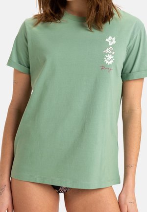 Femme portant une chemise vert sauge à manches courtes avec un revers roulé et des motifs tropicaux blancs sur la poitrine gauche.