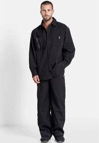 Hombre con pelo corto y barba, vistiendo una camisa negra holgada y texturizada y pantalones, con gafas de sol en el bolsillo de la camisa, de pie frente a un fondo blanco.