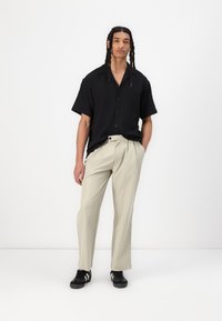 Maglietta nera a maniche corte con un motivo testurizzato, pantaloni beige chiaro e scarpe nere con strisce bianche, in piedi davanti a uno sfondo neutro.