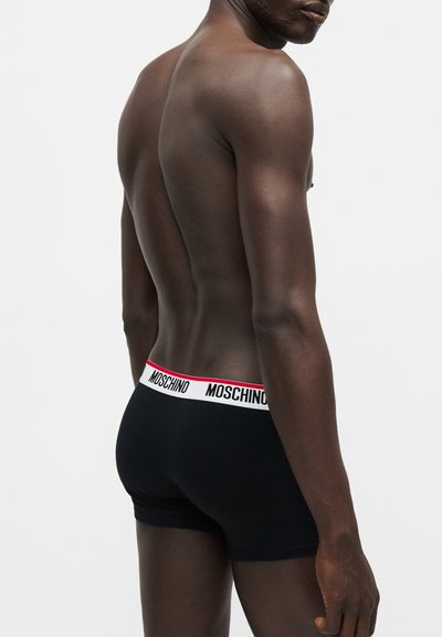 Moški od zadaj, oblečen v črne Moschino boksarice z belim pasom, rdečo obrobo in črno Moschino znamko.