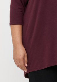 Túnica bordô com um corte descontraído, mangas três quartos e uma bainha curva. Combinada com leggings pretas, mostrando uma mão na bainha.
