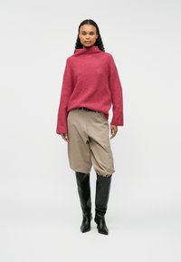 Liels rozā adīts džemperis ar augstu apkakli, bēšas platās bikses un melni ceļgala augstie zābaki ar papēžiem, uz balta fona.