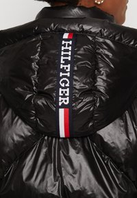 Doudoune noire avec une finition brillante, texture matelassée et une capuche ornée d'une sangle bleu marine affichant le logo et les couleurs "HILFIGER".