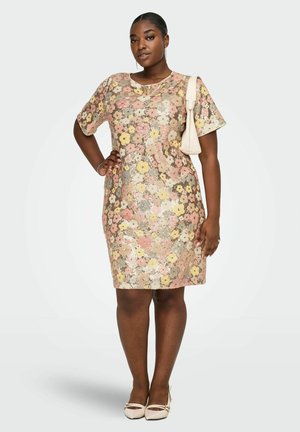 Kvinde iført en knælængde blomsterkjole i pastelnuancer, bærende en lille beige skuldertaske, stående med den ene hånd på hoften og et neutralt ansigtsudtryk.