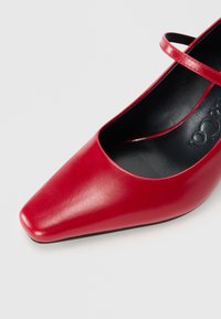 Scarpa Mary Jane rossa in pelle con punta quadrata e cinturino singolo sopra il collo del piede su sfondo bianco.