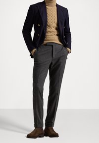 Granatowa marynarka dwurzędowa na beżowym swetrze w warkocze, zestawiona z szarymi, dopasowanymi spodniami i brązowymi zamszowymi botkami za kostkę.